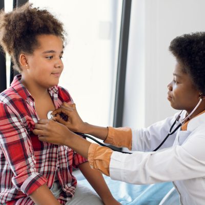 African female pediatrician with stethoscope auscultating cute teen mixed race girl patient. Black physician, checks heartbeat and lungs breathing of kid, making checkup before vaccination