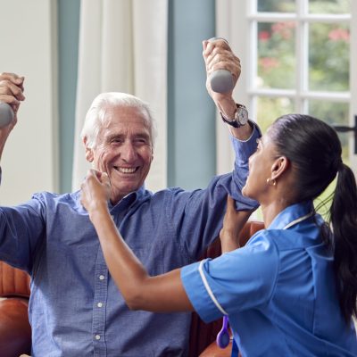 Female Nurse Or Physiotherapist In Uniform Helping Senior Man To Lift Hand Weights At Home
